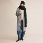 Person wearing a long gray coat, black beanie, green scarf, beige sweater, blue jeans, and black shoes on a beige background.