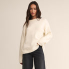 Woman wearing a cream sweater and dark pants against a plain background
