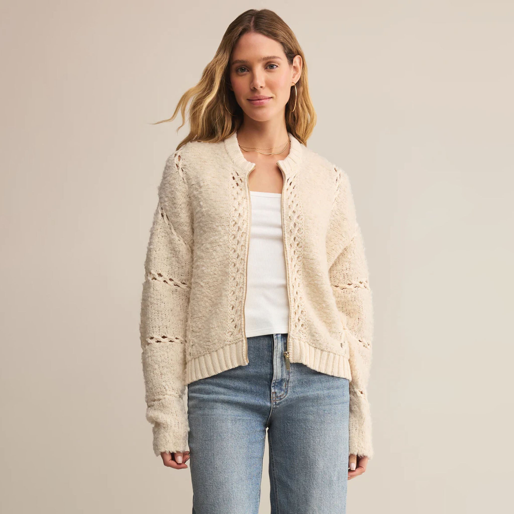 Woman wearing Z Supply Sundance Sweater Jacket in Crystal, a beige cardigan over a white shirt and blue jeans on a plain background