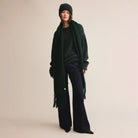 Woman wearing green sweater with a green beanie, green scarf, and dark pants on a plain background.