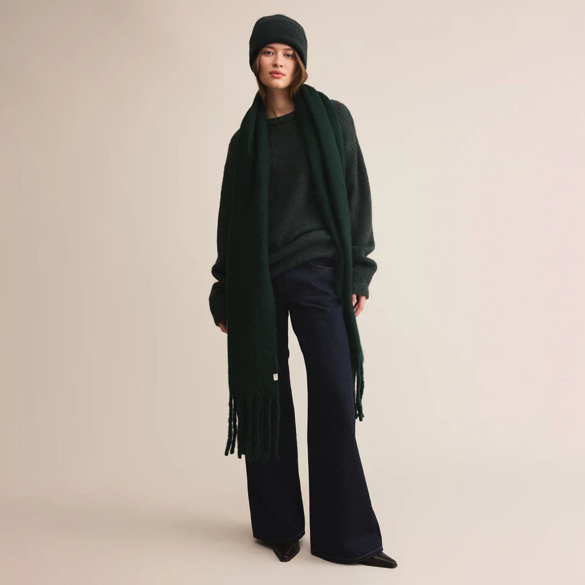 Woman wearing green sweater with a green beanie, green scarf, and dark pants on a plain background.