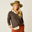 Woman wearing a brown sweater, white shirt, and red pants with a green hat on a plain background