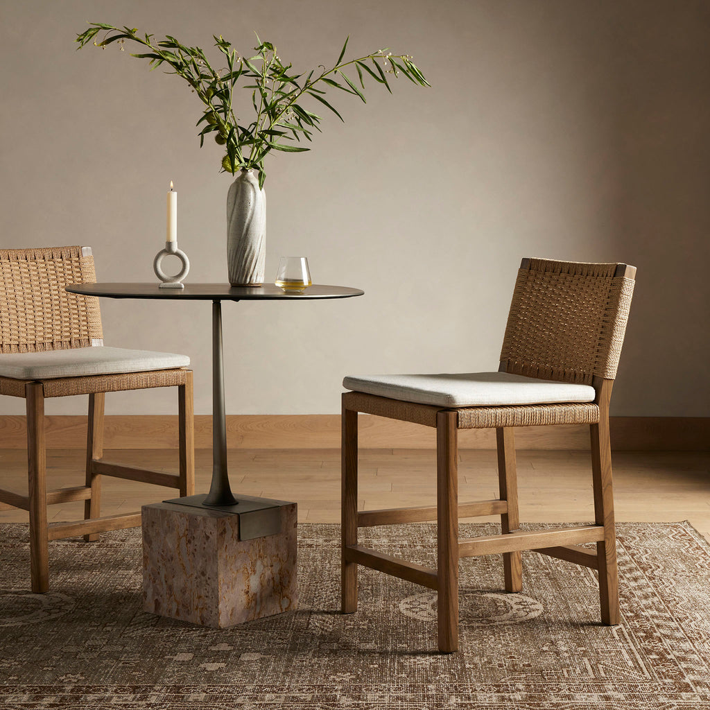 Four Hands Zuma counter stool with beige cushion and woven back in Dune Ash on a tribal rug in front of a round pedestal table