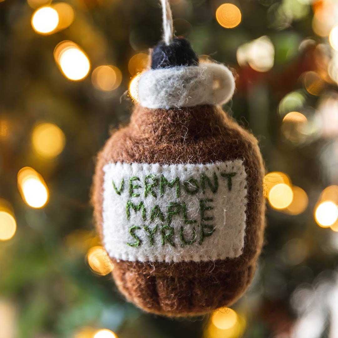 Farmhouse Pottery Felt Vermont Maple Syrup Ornament with a Christmas tree in the background at Addison West