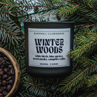 A dark glass candle jar labeled 'Winter Woods' by Burnwell Illuminaries, surrounded by pine branches and coffee beans, with a wooden bowl of coffee beans at the lower left.