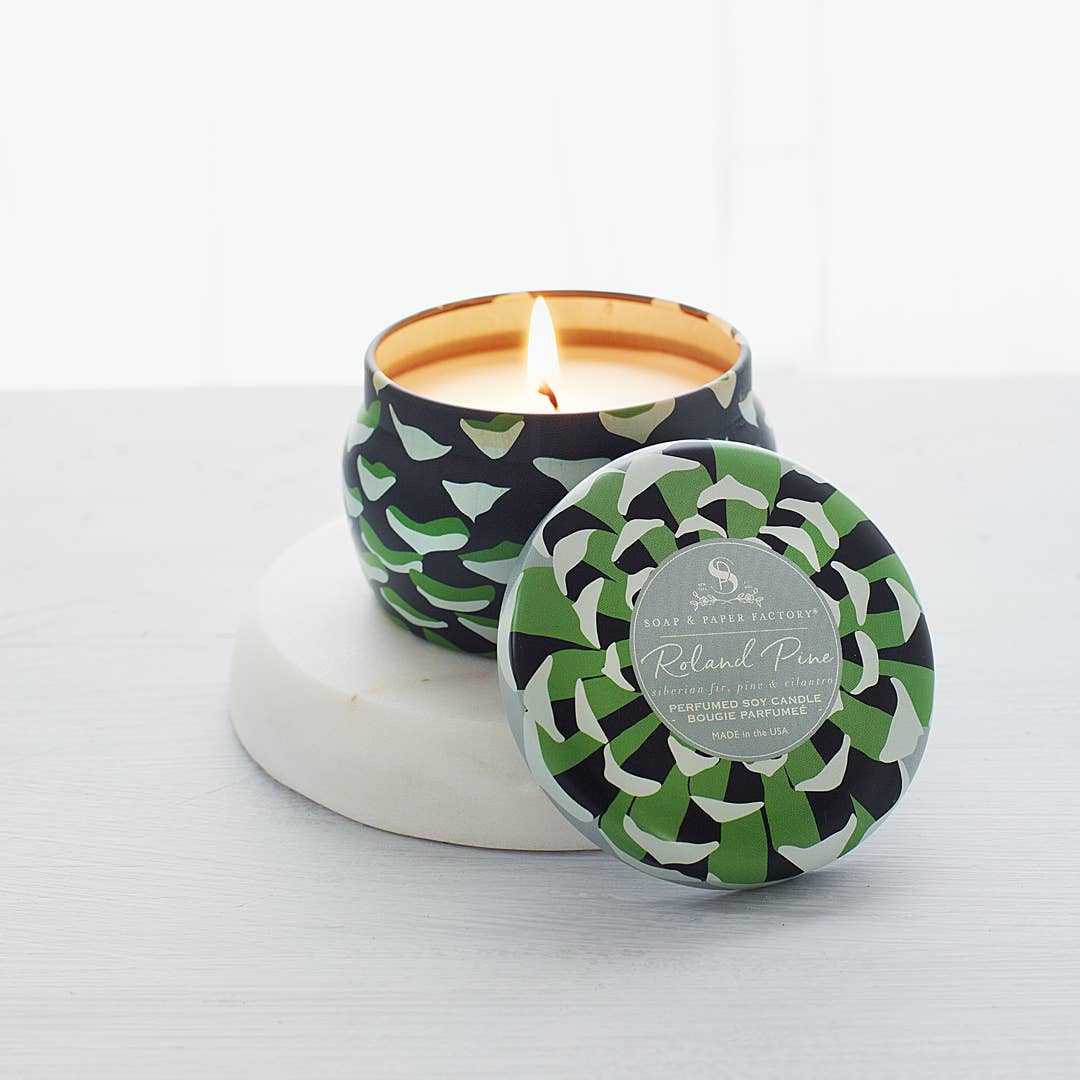 A small Roland Pine tin soy candle with a green and black geometric pattern lid, burning with a visible wick, on a white surface.