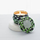 A small Roland Pine tin soy candle with a green and black geometric pattern lid, burning with a visible wick, on a white surface.