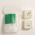 A clear resealable pouch labeled Forest by Wild Cover Rituals with a green label, shown next to two small square white bath salts with dried botanical flecks on a light beige background.