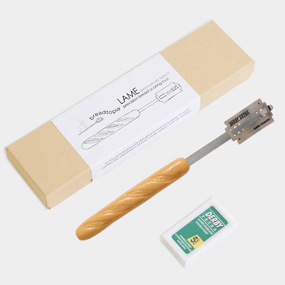Bread lame with wooden handle and metal blade, next to a loaf of bread and a box labeled 'LAME'.