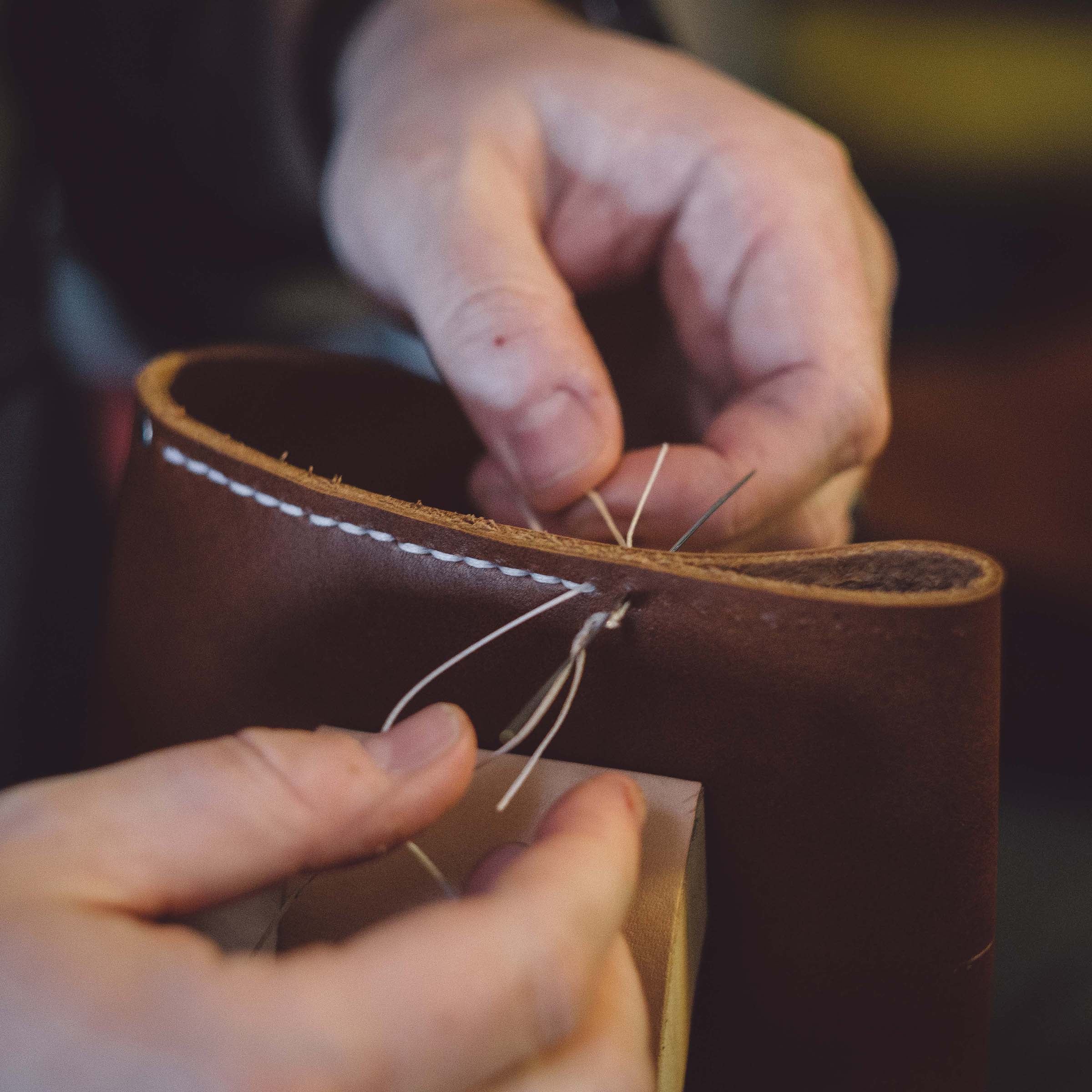 Close-up of a person's hands stitching a brown leather item, showing white stitching and the texture of the leather.