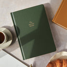 Green recipe book on a table with a cup of tea and croissant