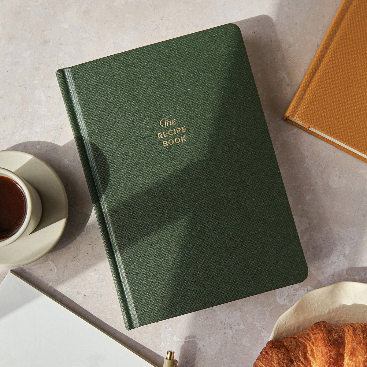 Green recipe book on a table with a cup of tea and croissant