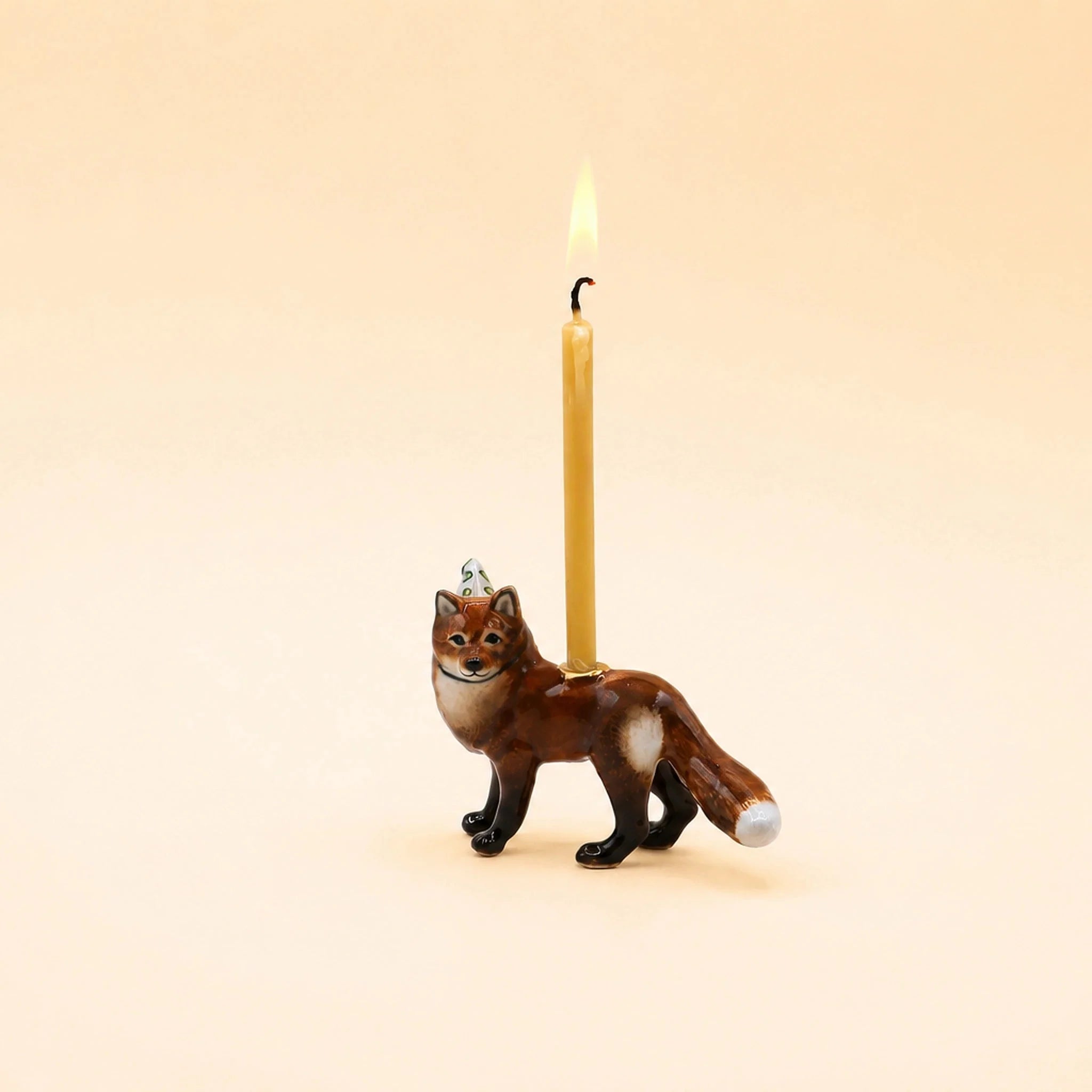 A small ceramic fox candle holder with a lit yellow taper candle inserted behind it, set against a solid warm beige background.