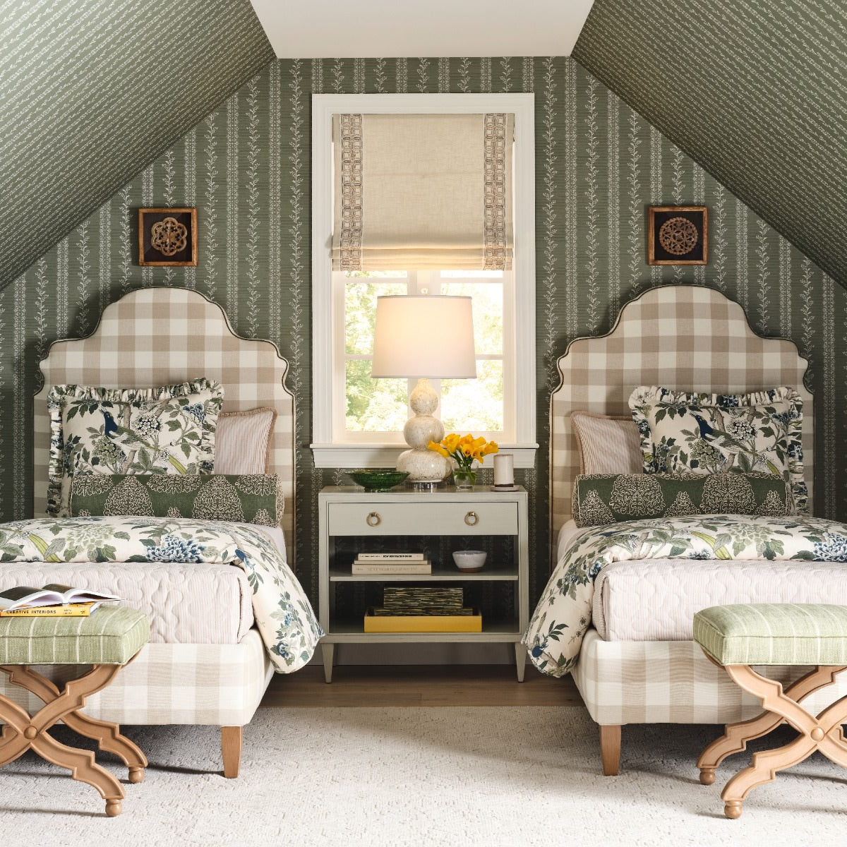 A symmetrical bedroom setup with two single beds facing each other under a slanted ceiling, featuring green patterned wallpaper, beige-and-white gingham headboards, floral bedding, matching green-and-beige ottomans, a small nightstand with a lamp, and framed wall art.