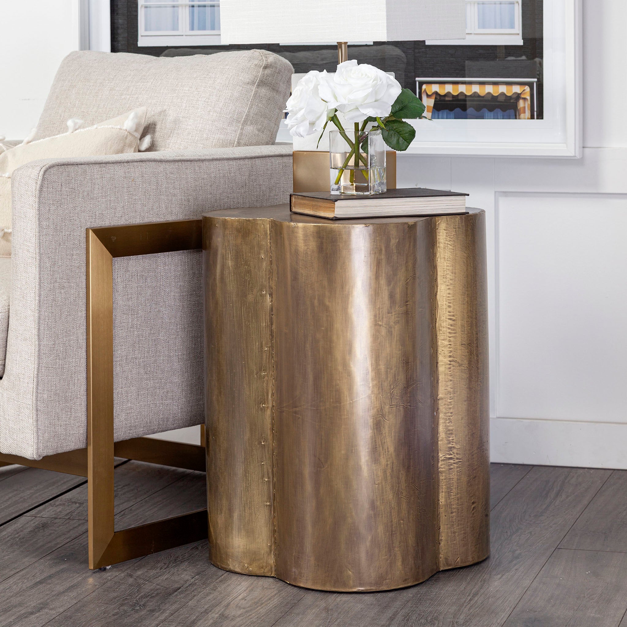 Clover shaped side table with brass cladding on a white background beside a white sofa with a vase of white flowers.