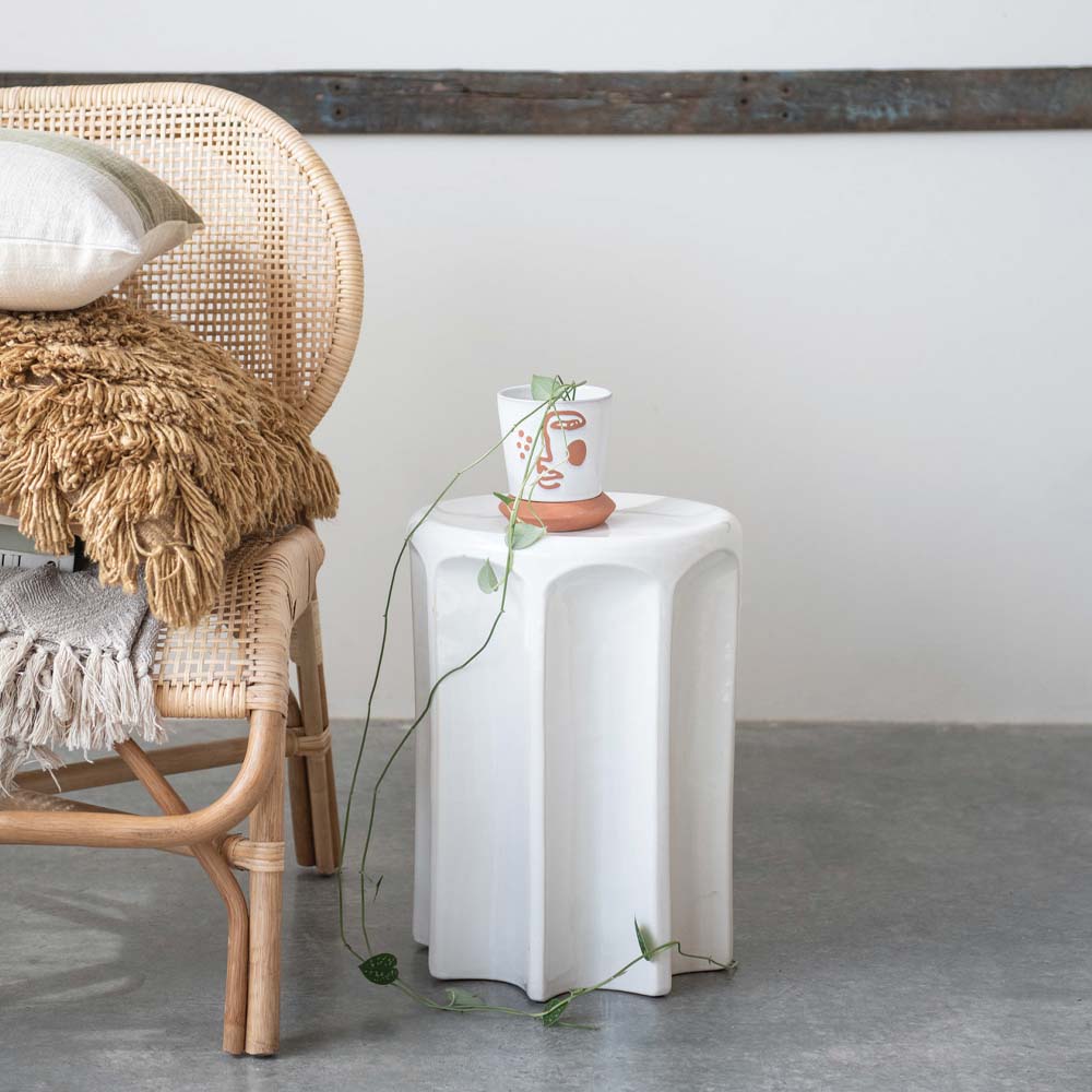 Cane chair on concrete floor with pillows and blankets stacked up next to white stoneware stool