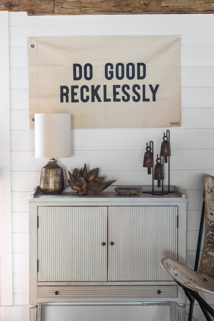 White washed wood cabinet with lamp and flag above that says Do Good Recklessly hanging above 