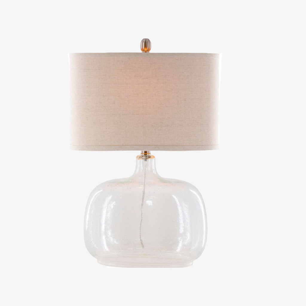 Surya brand Bentley table lamp with glass base and beige linen shade on a white background