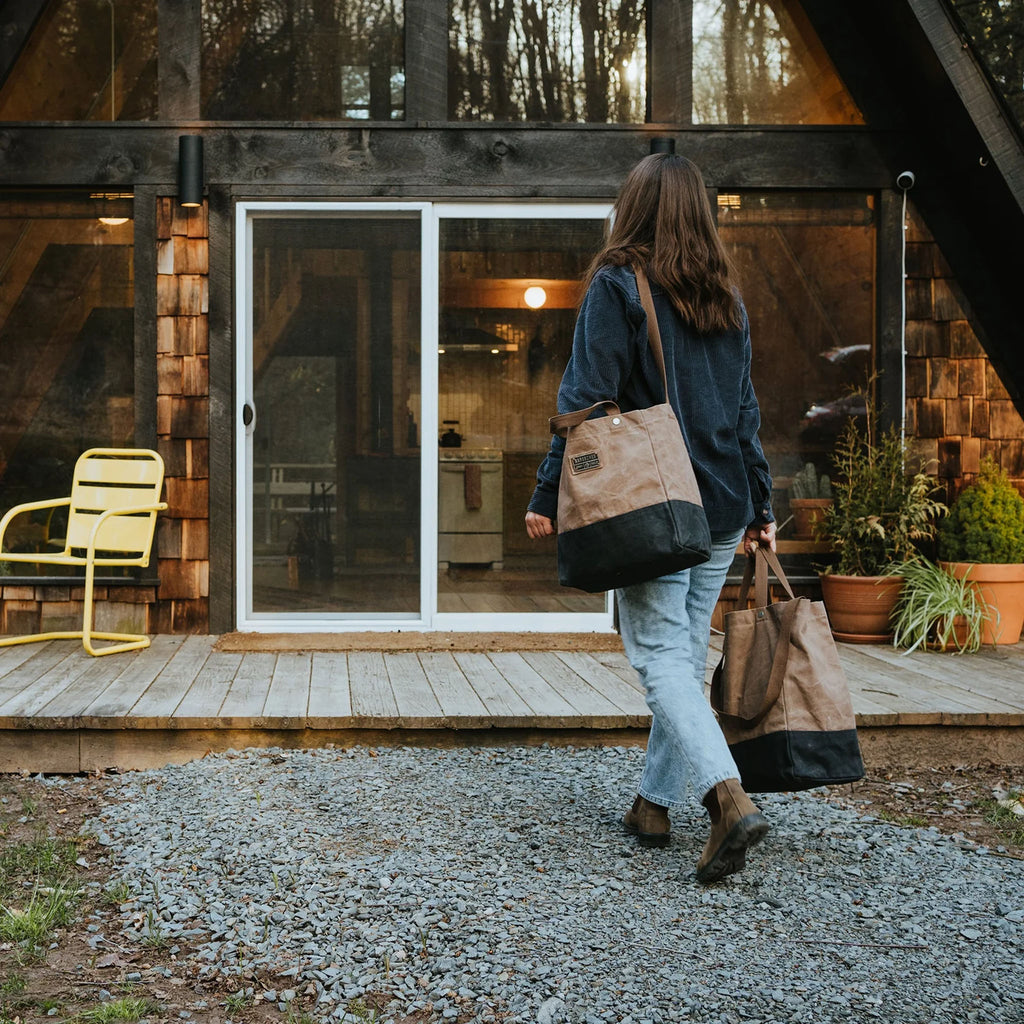 Woman carrying two bare bones neelum totate bags on a gravel walk way into an A frame house