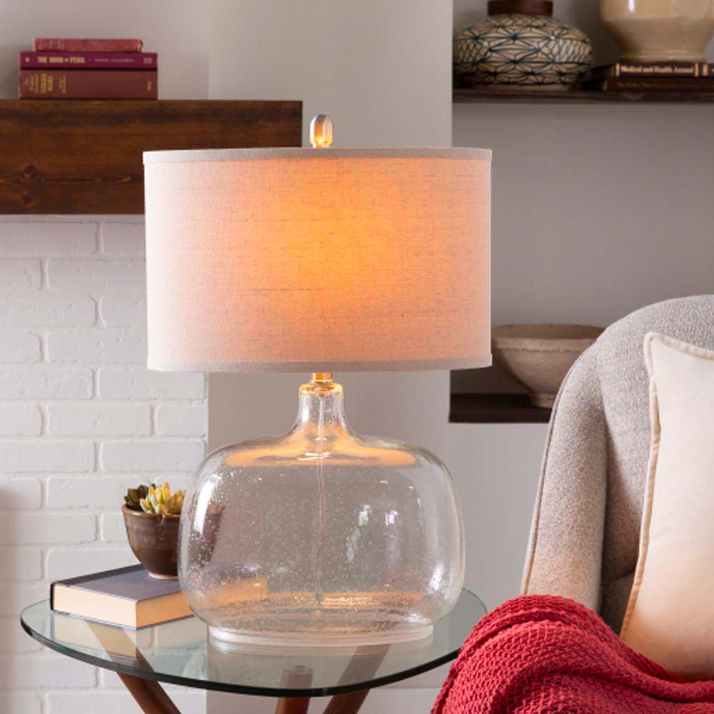 Surya brand Bentley table lamp with glass base and beige linen shade on a side table next to a chair and white brick fireplace