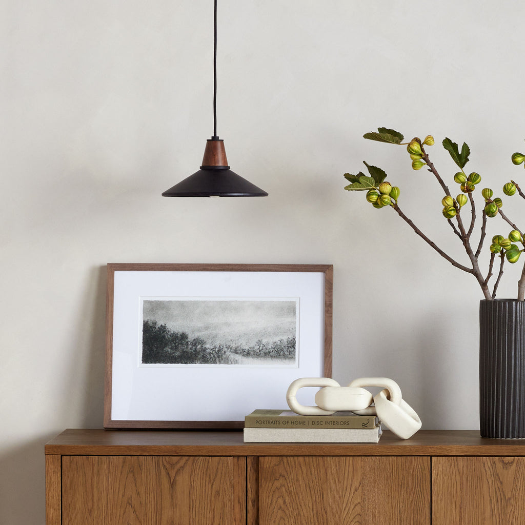 Black leather and walnut pendant light with black cord above wood sideboard with picture leaning and large black vase with greenery