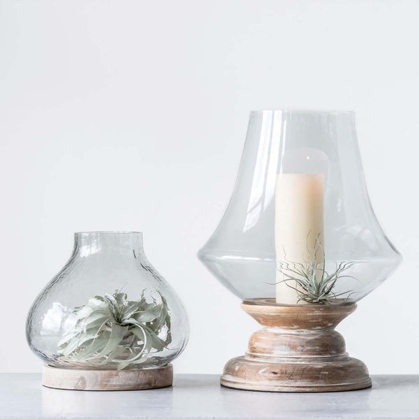 Two large handblown glass and wood hurricanes with candles and plants on a white background