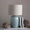 Blue green creative coop brand stoneware lamp with linen shade on a shelf with books white background