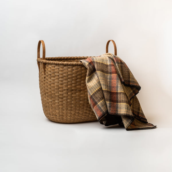 Large handmade rattan basket with arched handles and wool blanket draped over the side on a white background