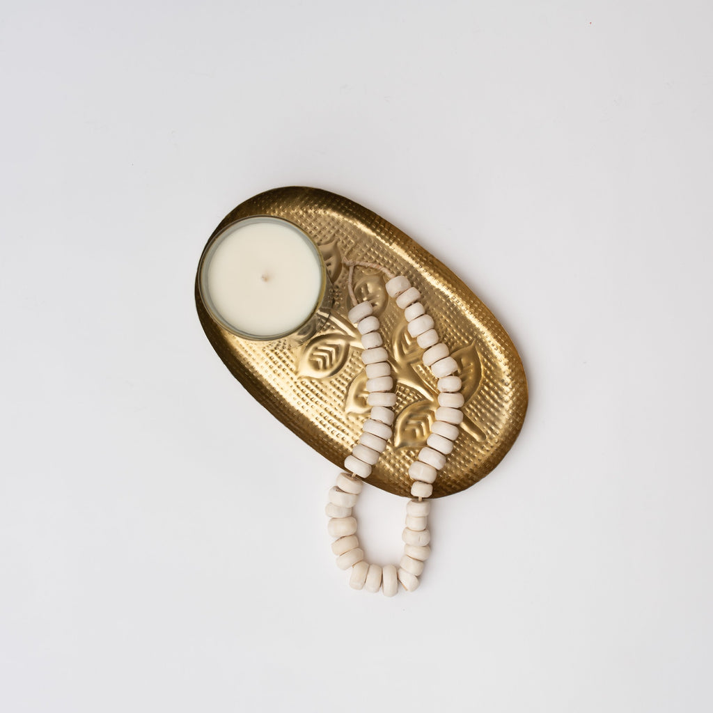 Embossed brass colored iron tray with leaf pattern with candle and beads on a white background