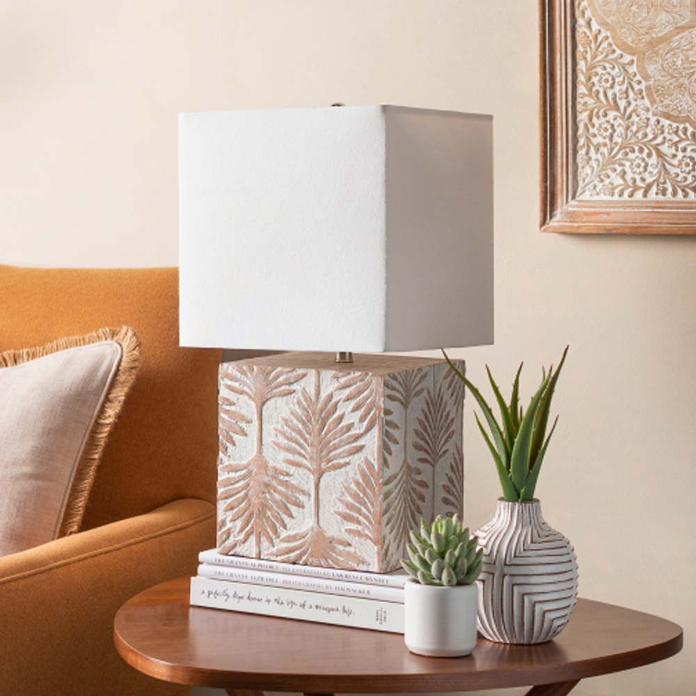 Surya brand Dax table lamp with whitewashed square wood base with leaf pattern and square white shade on a side table with books and plants 