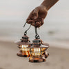 Person's hand holding three Barebones brand mini Edison lantern with vintage style and caged bulb in copper on a beach