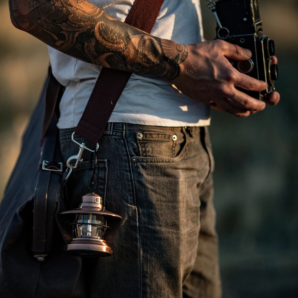 Man with tattoos holding camera and bag with Barebones brand mini Edison lantern with vintage style and caged bulb in copper hooked to bag