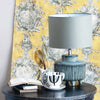 Blue stoneware table lamp with footed base and khaki green linen Shade on a table with yellow wallpaper in the background