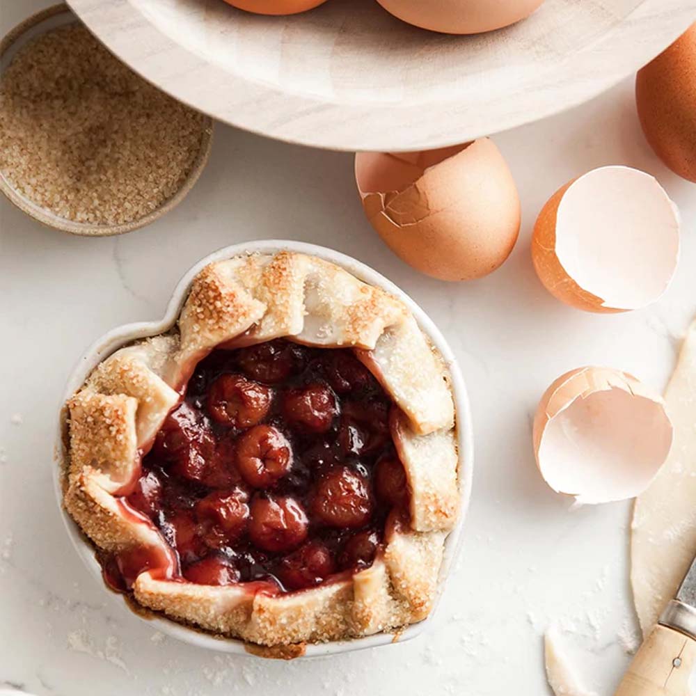 Farmhouse pottery brand heart dish filled with cherry pie on a counter with eggs and baking products