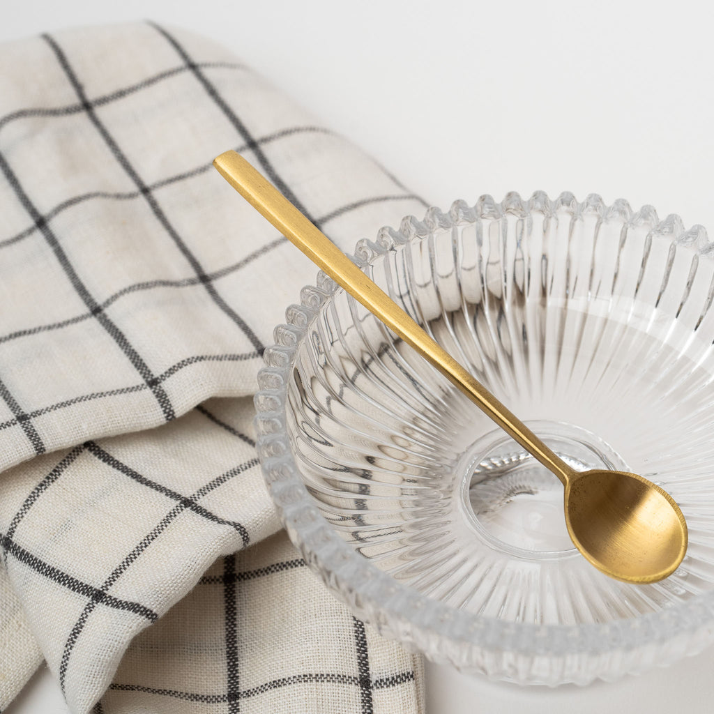 Pressed glass footed bowl with brass spoon and checked napkin on a white background