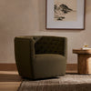 Barrel style 'Hanover' swivel chair upholstered in olive performance fabric by Four Hands furniture in a living room with patterned rug beside table  with books and wine glass