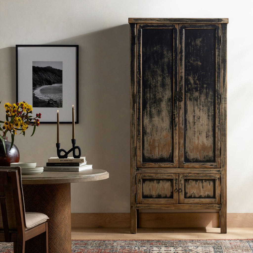 Tall 'Hitchens' cabinet with worn black paint and two doors in a dining room with rug and table with books and candles 