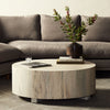 'Hudson' round coffee table by four hands furniture in light bleached spalted wood color in a living room beside dark grey couch 