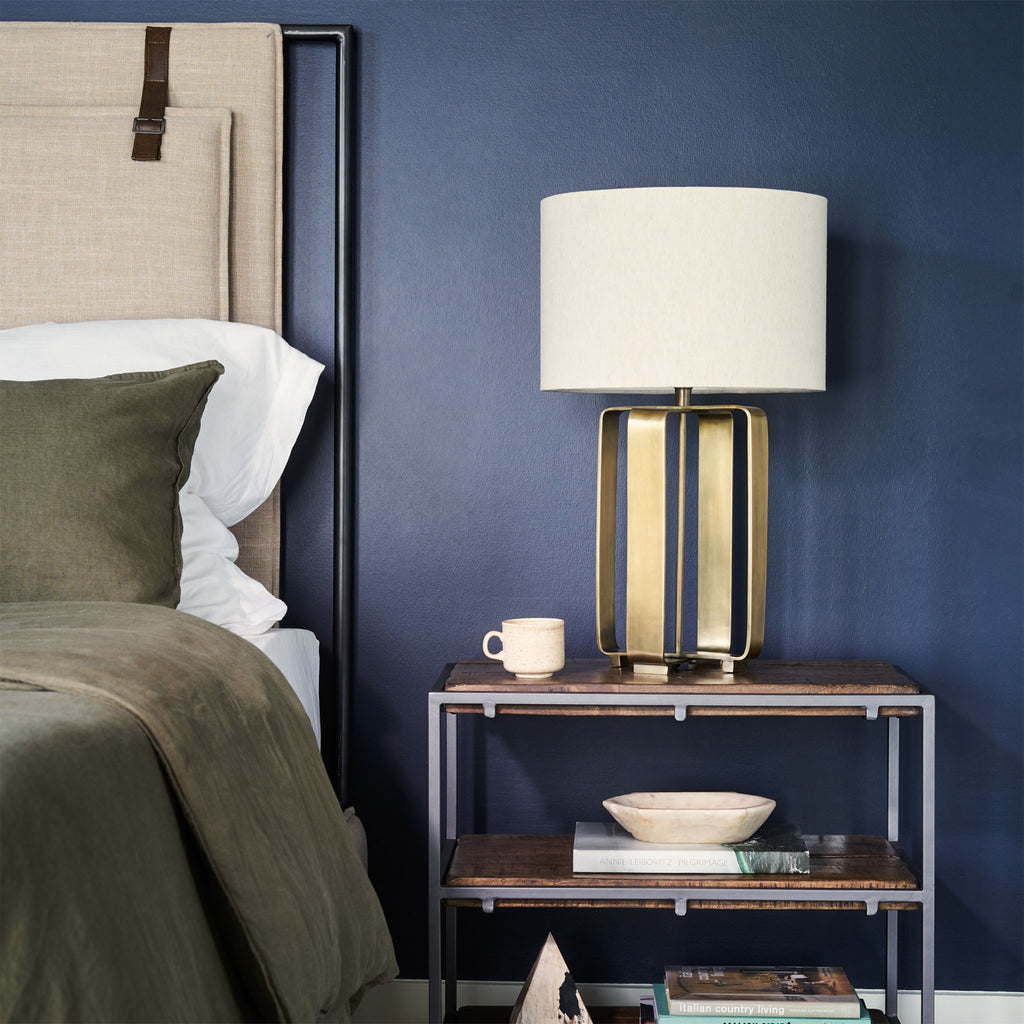 Upholstered 'Leigh' bed by Four hands furniture with metal frame and buckled head board panels in navy blue painted bedroom with three tier night stand and brass lamp