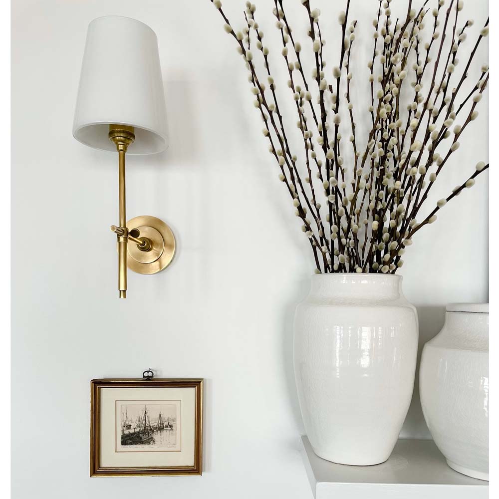 Set of two white ginger jars on a mantle beside a brass sconce and painting.