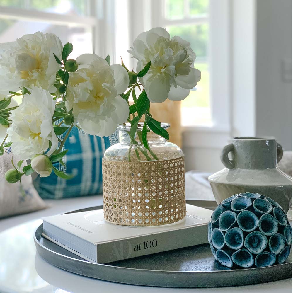 Cane wrapped vase filled with white peonies on a table with book and decorative accessories