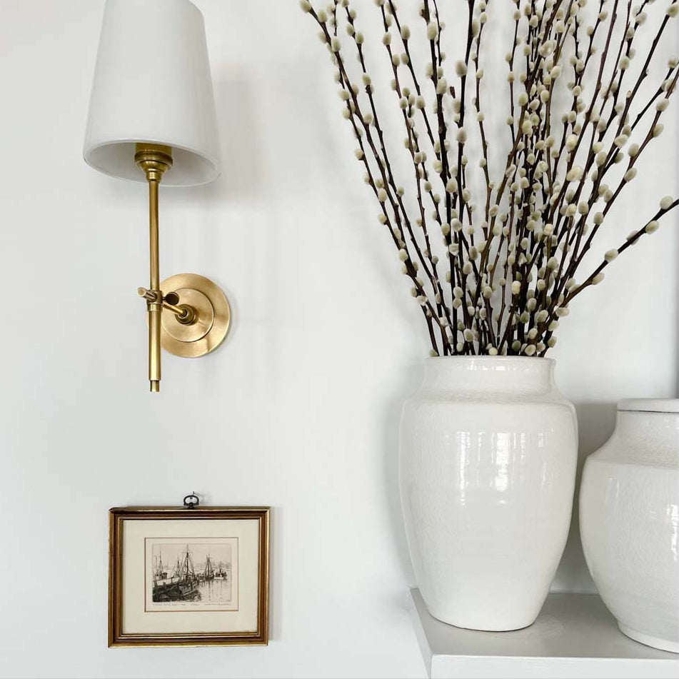 Set of two white ginger jars on a mantle beside a brass sconce and painting. 