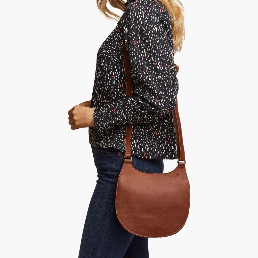 Model wearing Able brand Martha saddle bag in whiskey color with adjustable shoulder strap on a white background