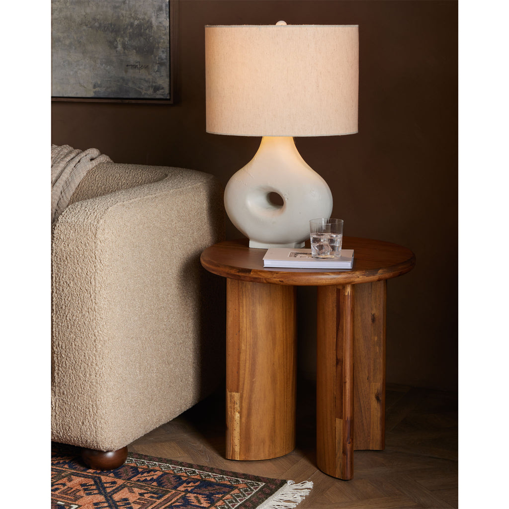 Living room with sofa and rug and white lamp sitting on Stained acacia wood round end table with two rounded legs by four hands furniture 