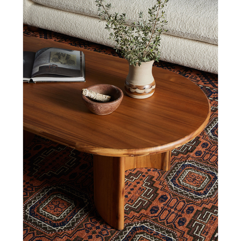 Four hands brand oval wood 'Paden' coffee table with vase and book on a tribal patterned rug next to cream colored sofa
