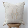 Natural square throw pillow with hand embroidered geometric pattern placed on a stool in front of white wall
