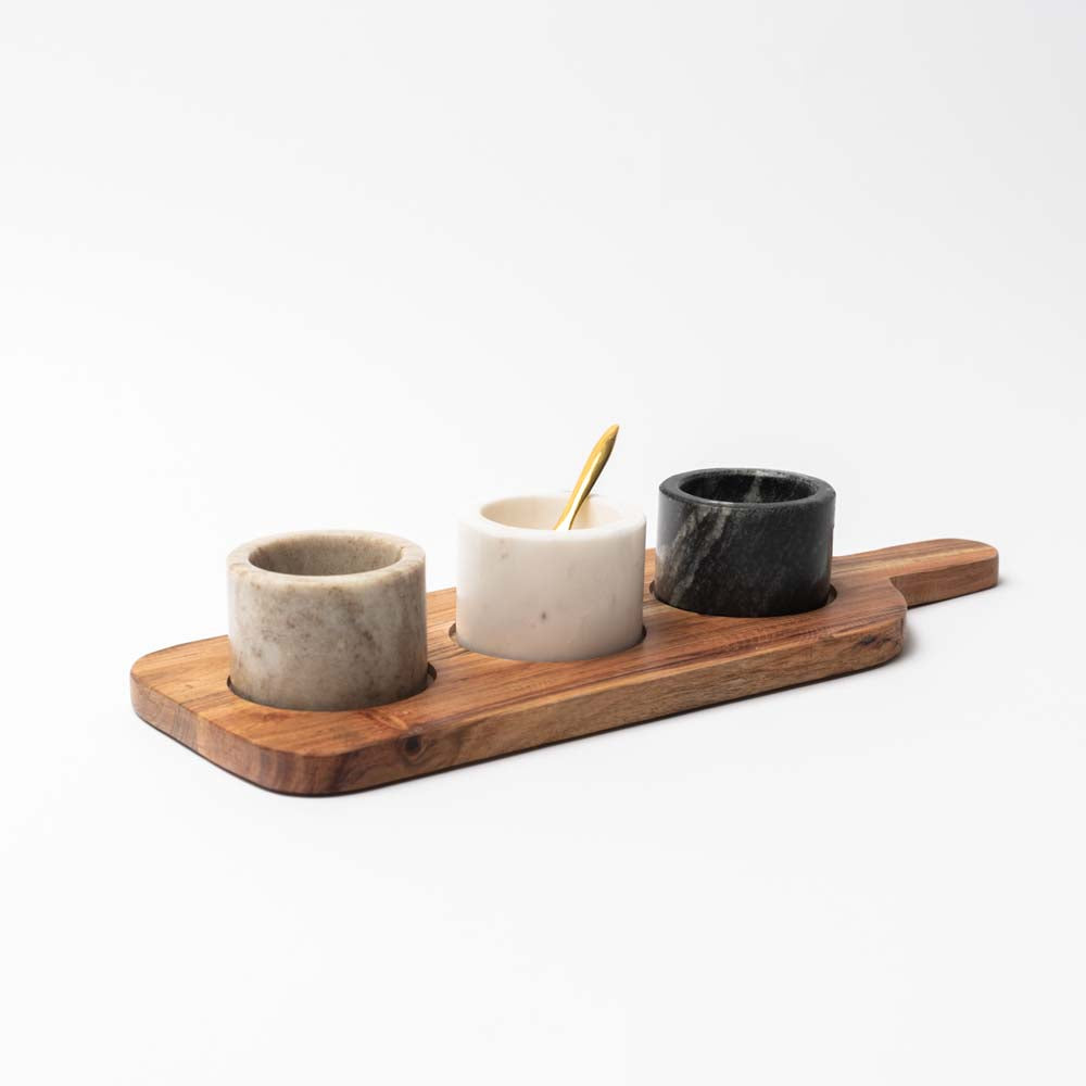 Wood serving board with three marble pinch pots and a brass spoon on a white background