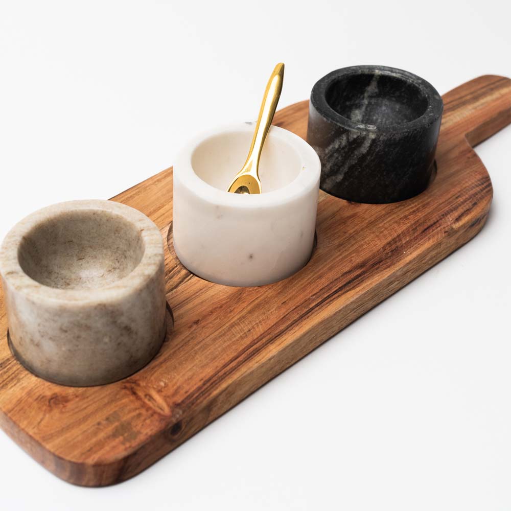 Wood serving board with three marble pinch pots and a brass spoon on a white background