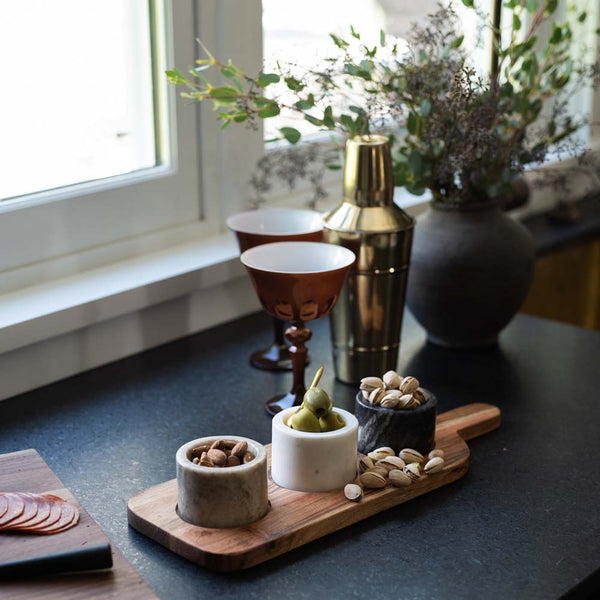 Wooden serving board with three small marble bowls and nuts on a counter with cocktail shaker in the background