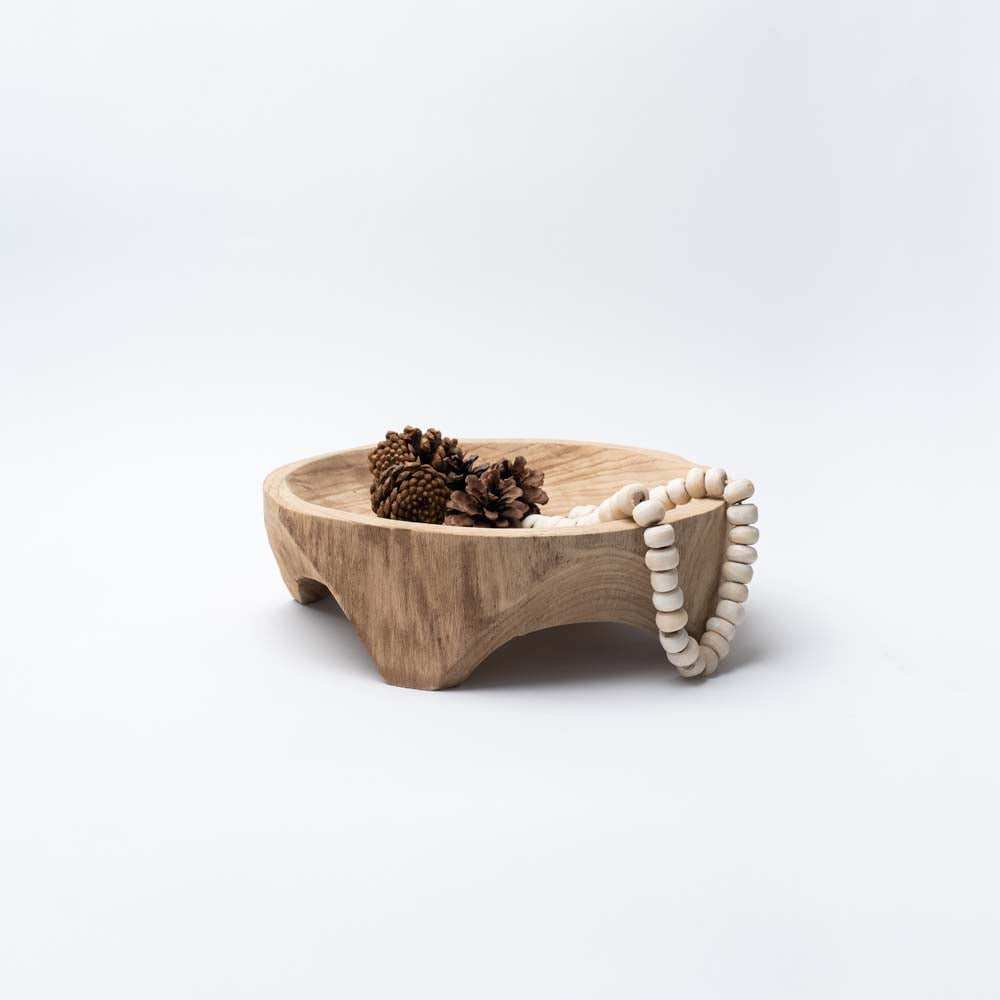 Shallow hand carved wood bowl with four feet filled with pinecones and beads on a white background
